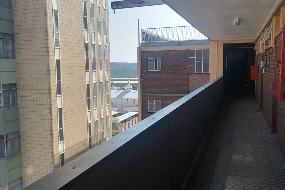 A covered exterior balcony walkway featuring tiled flooring and a metal balustrade, overlooking adjacent urban buildings.