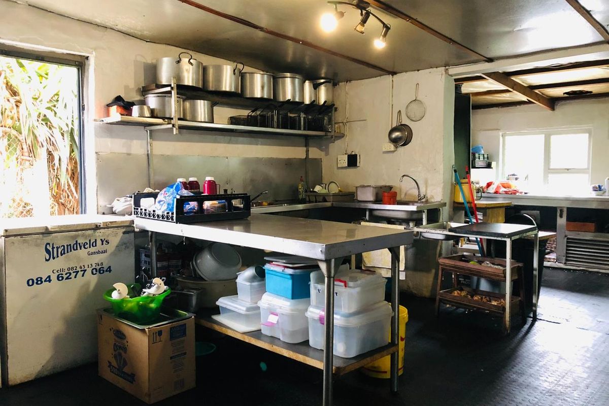Commercial kitchen space featuring stainless steel prep tables, industrial shelving, and a sink, with functional but dated finishes throughout.