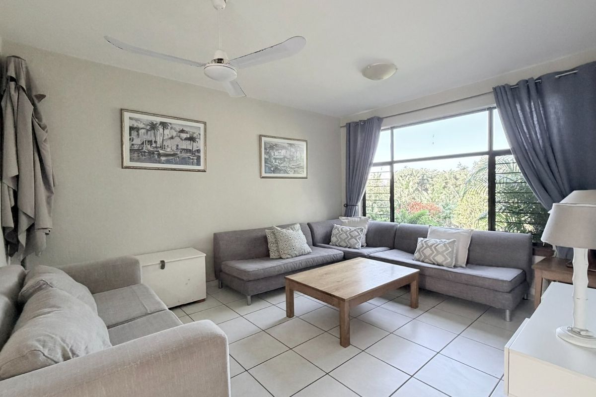 A bright living room featuring large windows with natural light, tiled flooring, and a ceiling fan, offering a comfortable and functional space.