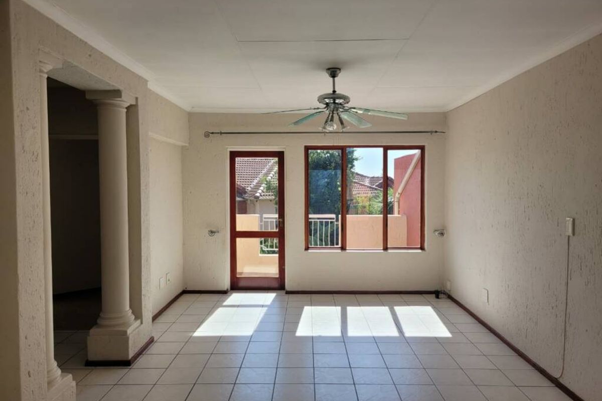An empty living room featuring tiled flooring, decorative columns, a ceiling fan, and natural light through timber-framed windows and a door.