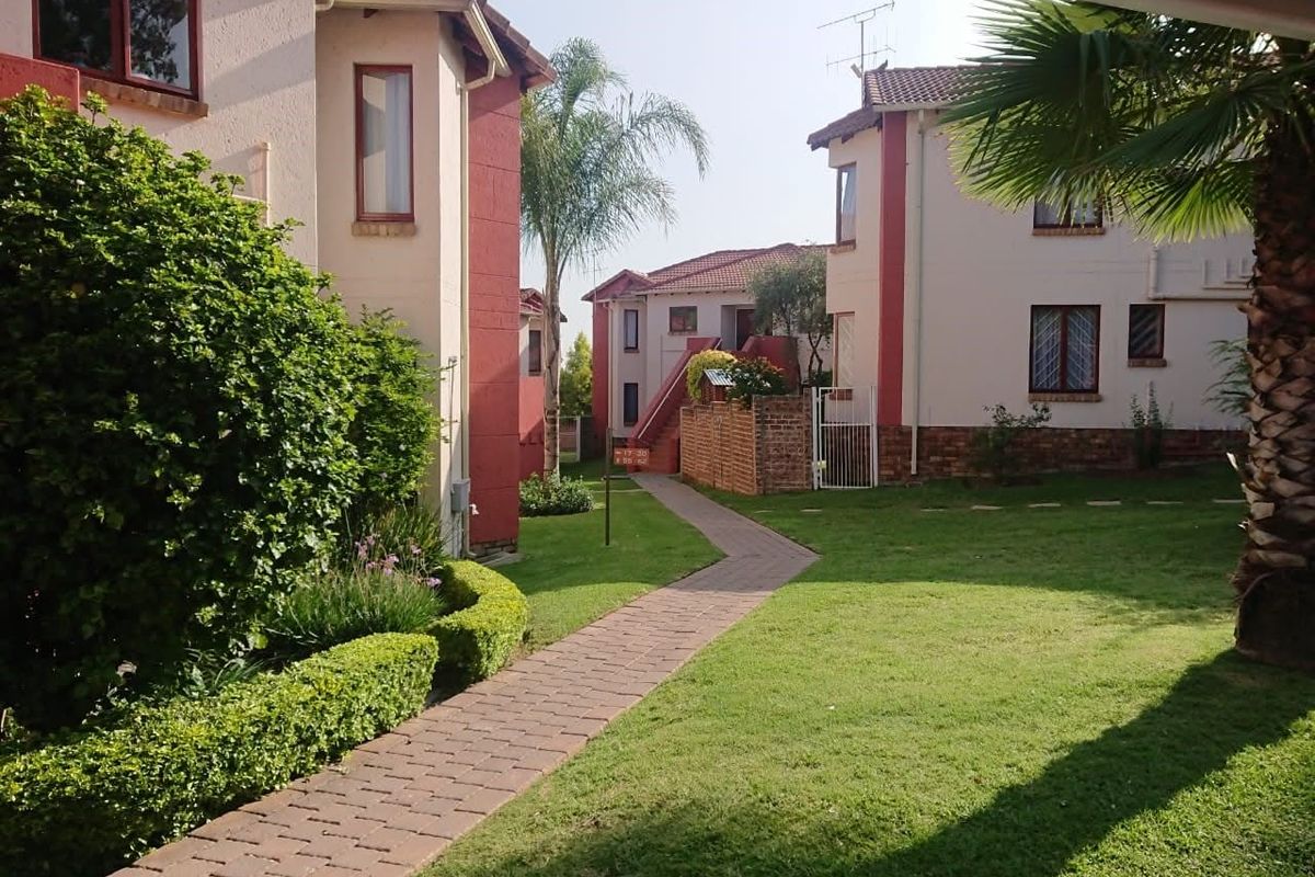 A view of the apartment complex grounds featuring a paved walkway, well-maintained lawn, and landscaped garden areas between the residential buildings.