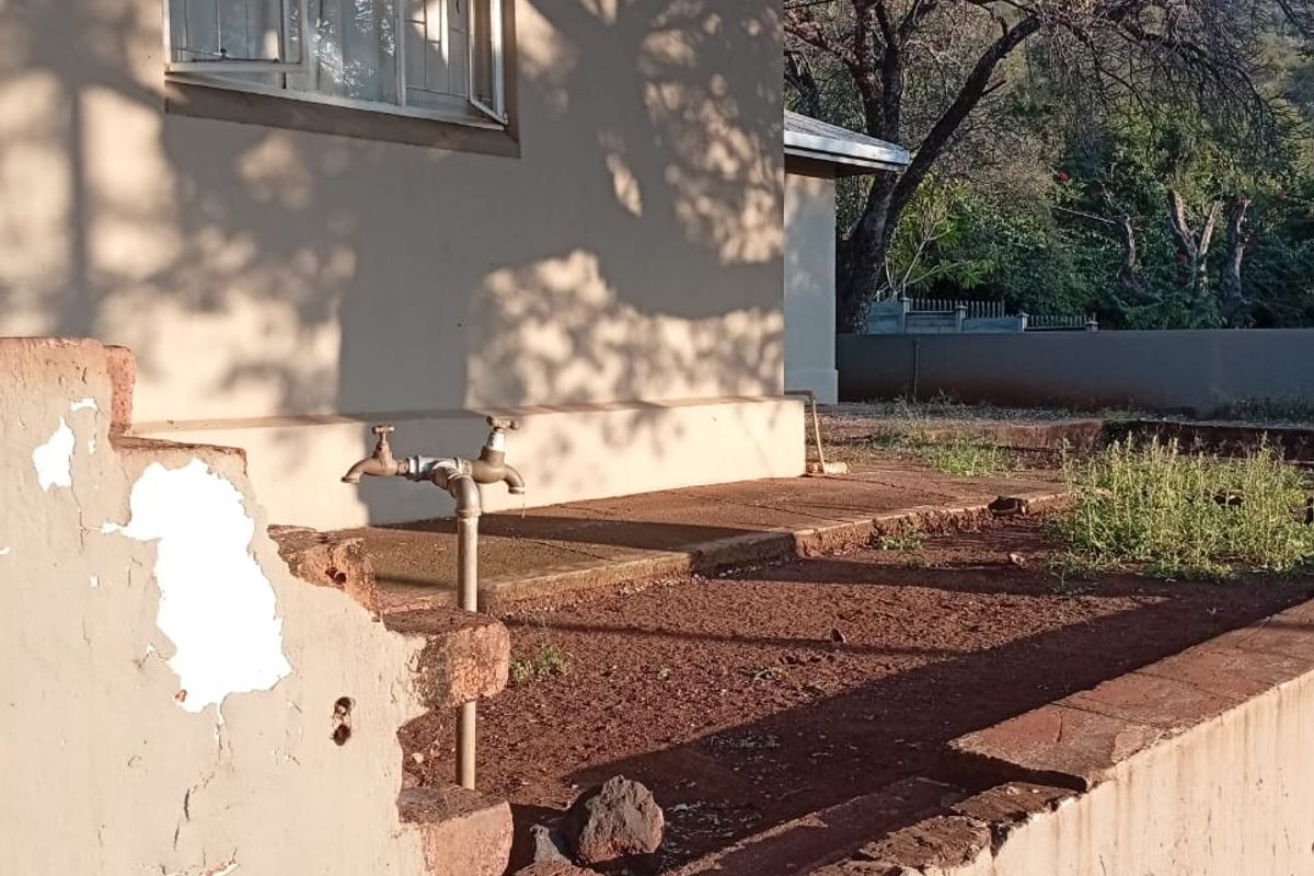Rear exterior view showing a concrete paved area, external water tap, and a damaged brick retaining wall requiring maintenance and repair.
