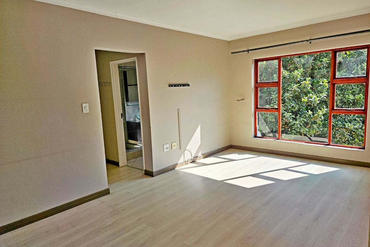 A bright, empty bedroom featuring light-toned laminate flooring, large windows with garden views, and direct access to an adjoining bathroom.