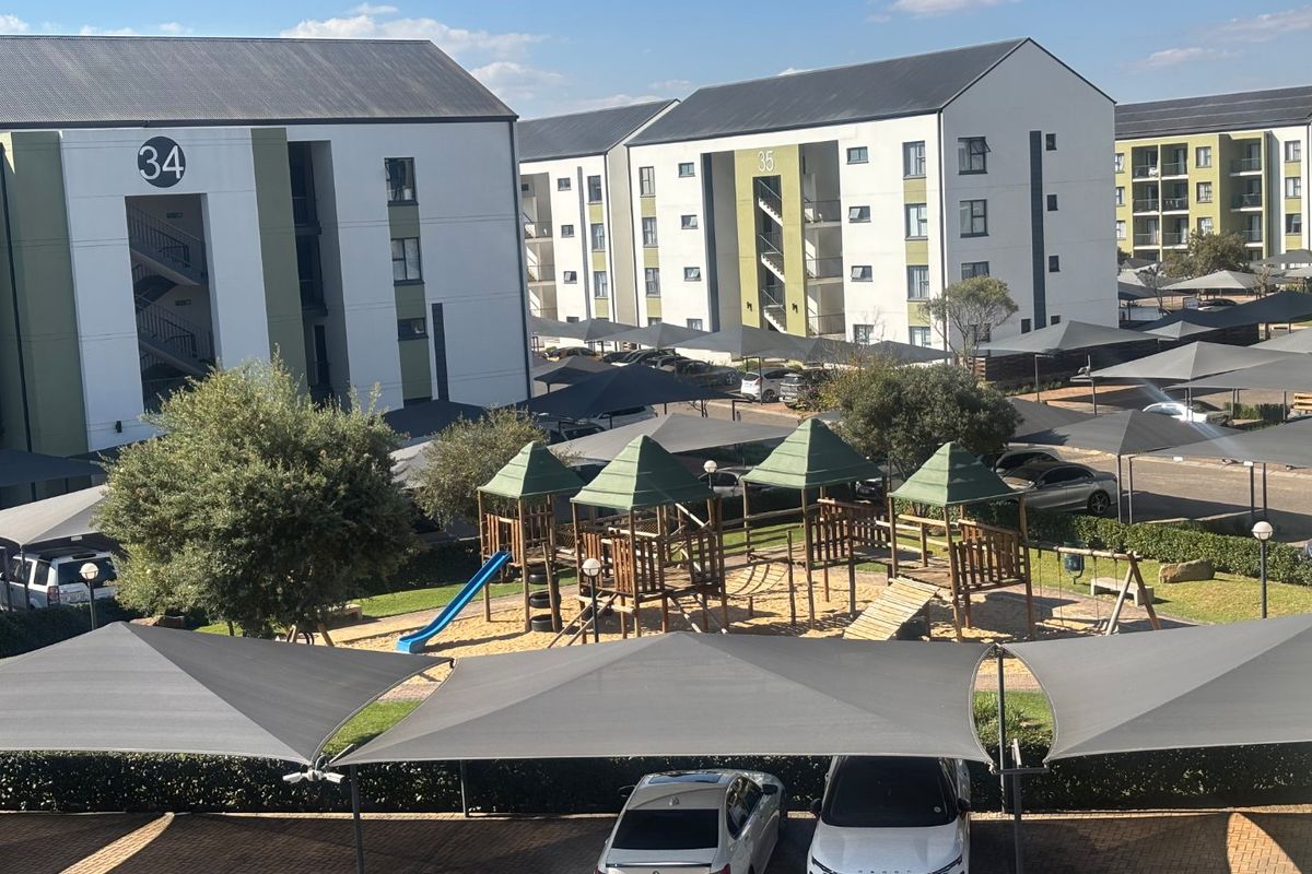 View of the apartment complex grounds featuring paved parking with shade cloth carports and a communal children's playground area.