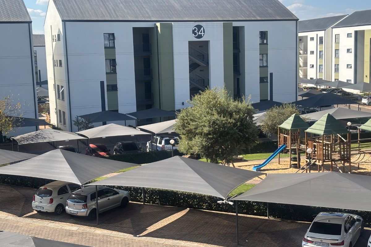 Aerial view of an apartment complex featuring paved parking with shade cloth carports, a communal playground, and multi-story residential buildings.