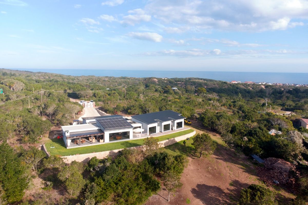 Aerial view of a modern farm property featuring a large residence with solar panels, a covered patio, and expansive surrounding landscape with ocean views.