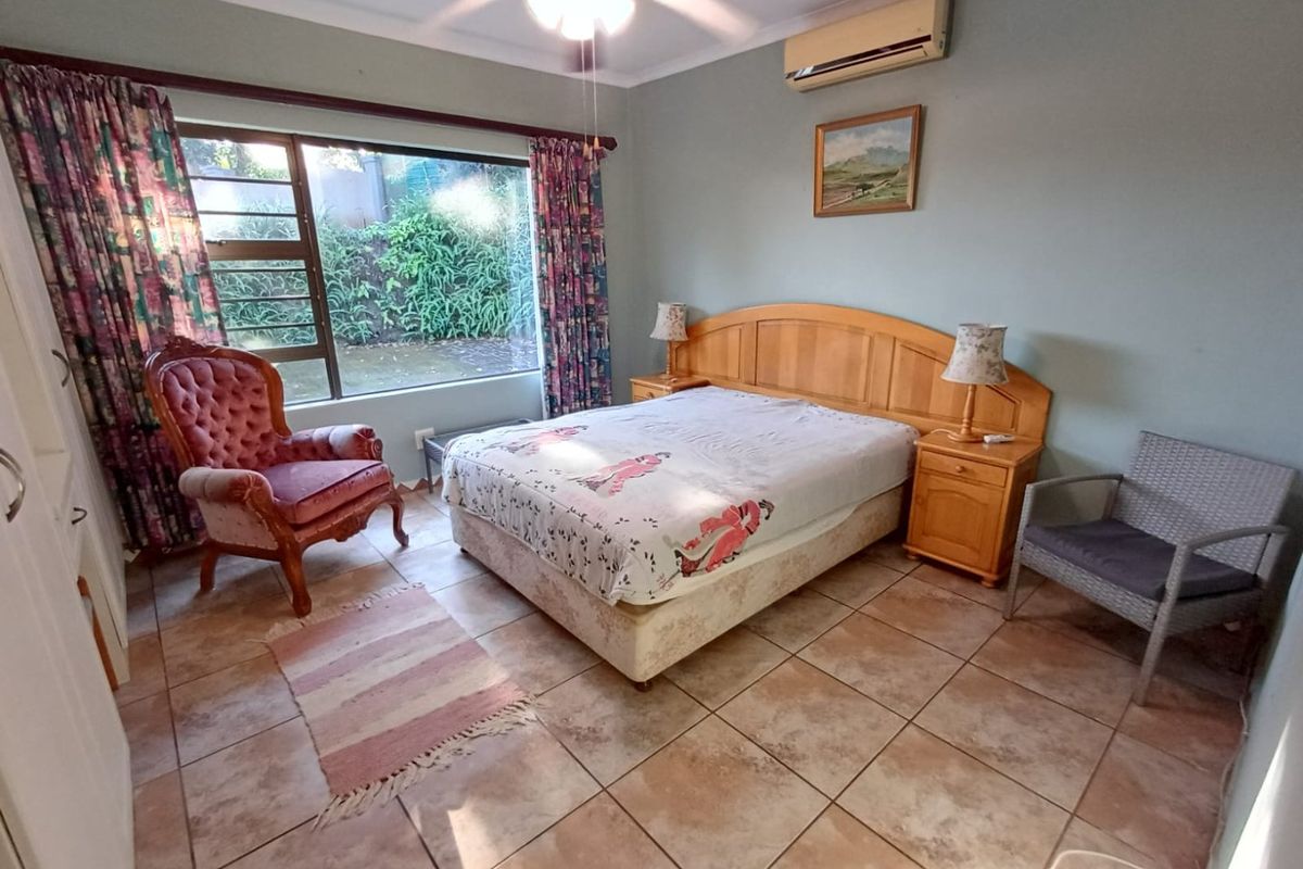 A spacious bedroom featuring tiled flooring, a ceiling fan, and a wall-mounted air conditioning unit, with large windows providing natural light.