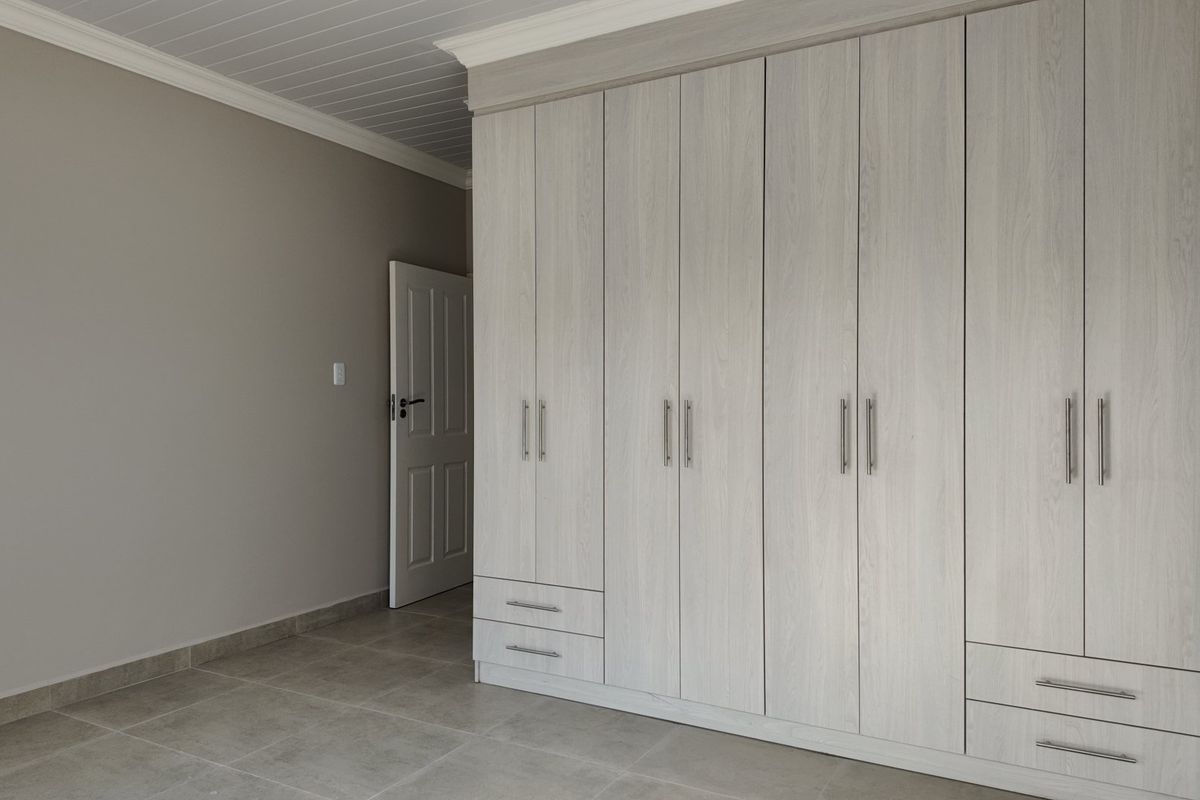 A clean, modern bedroom featuring extensive built-in cupboards with light wood-grain finish, neutral wall tones, and large format floor tiles.
