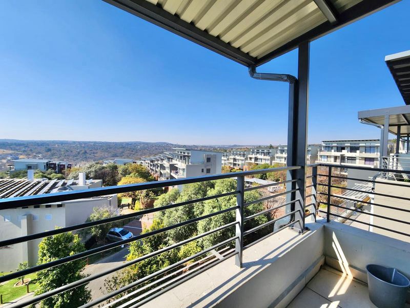 A covered balcony featuring a metal balustrade and tiled flooring, offering elevated views over the surrounding residential complex and landscape.