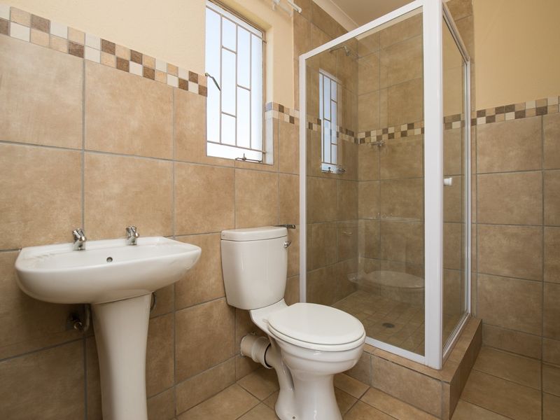 A functional bathroom featuring a pedestal basin, toilet, and a glass-enclosed shower with neutral floor-to-ceiling tiling and security bars on the window.