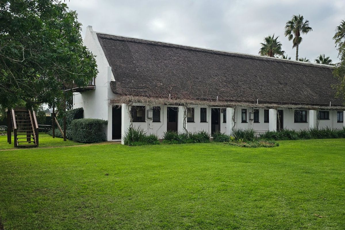 A rear view of a traditional building featuring a thatched roof, white facade, and an expansive green lawn with a timber staircase.