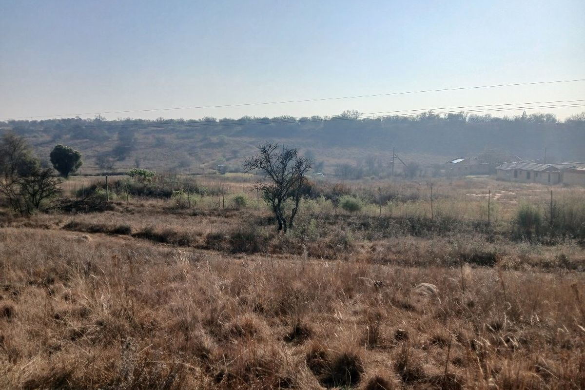 A wide-angle view of the vacant residential land featuring dry grass, sparse trees, wire fencing, and distant rural landscape.