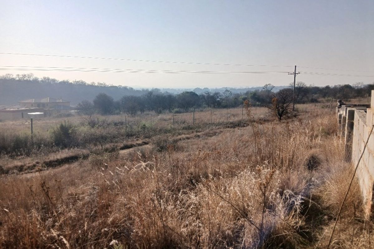 A wide-angle view of a vacant residential plot featuring dry vegetation, perimeter fencing, and distant landscape views.