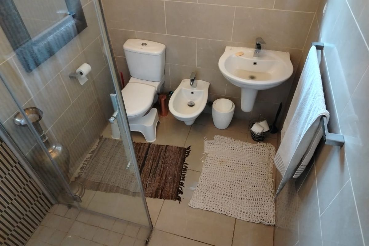 A compact bathroom featuring a pedestal washbasin, toilet, bidet, and a glass-enclosed shower with neutral ceramic wall and floor tiles.