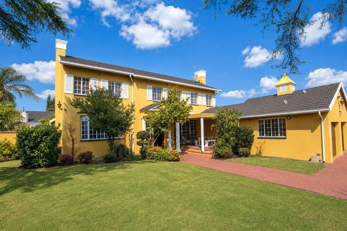 A well-maintained rear exterior view of a yellow two-story house featuring a paved walkway, covered patio, and landscaped garden.