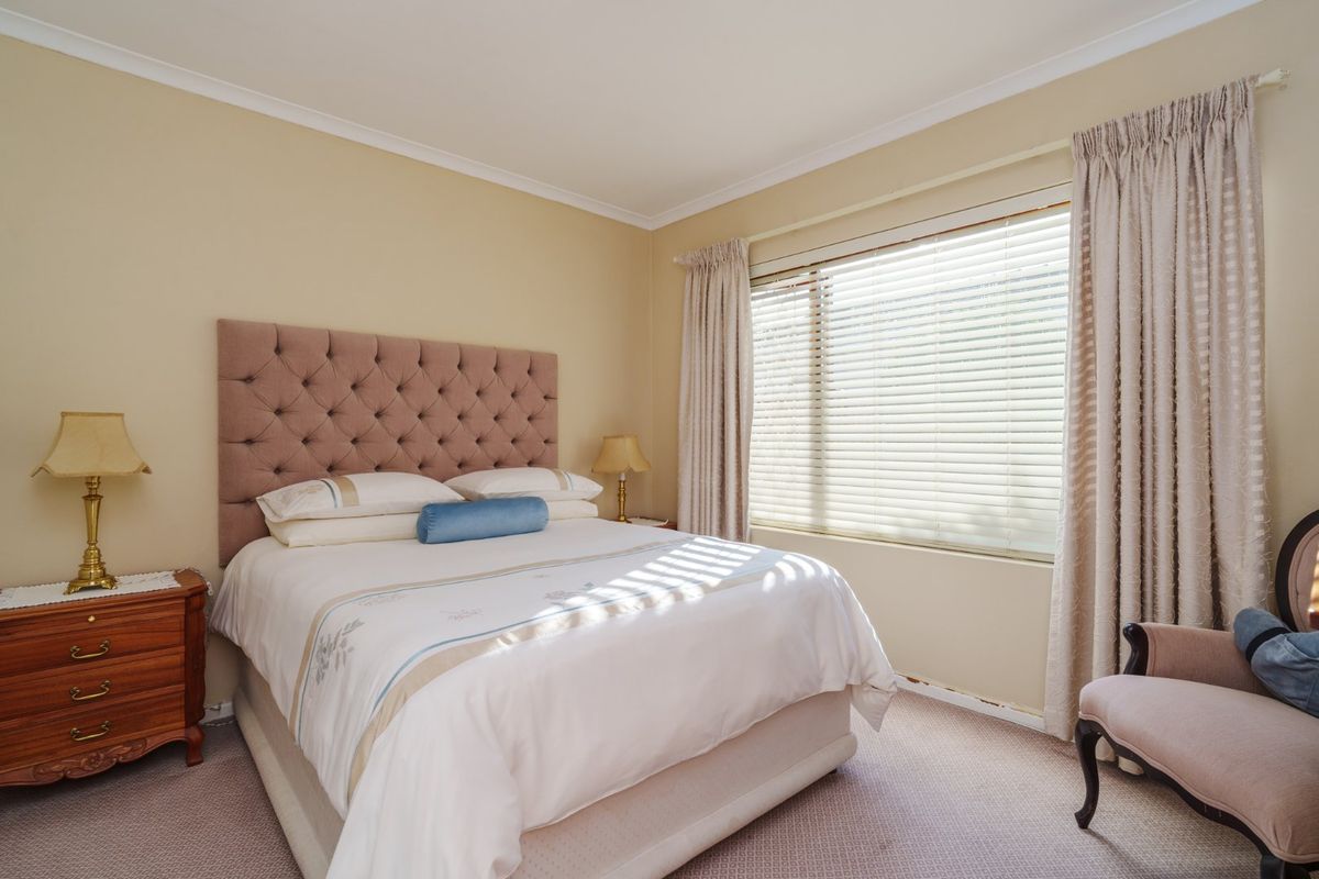 A well-lit bedroom featuring neutral wall tones, carpeted flooring, and large windows with fitted venetian blinds and curtains.