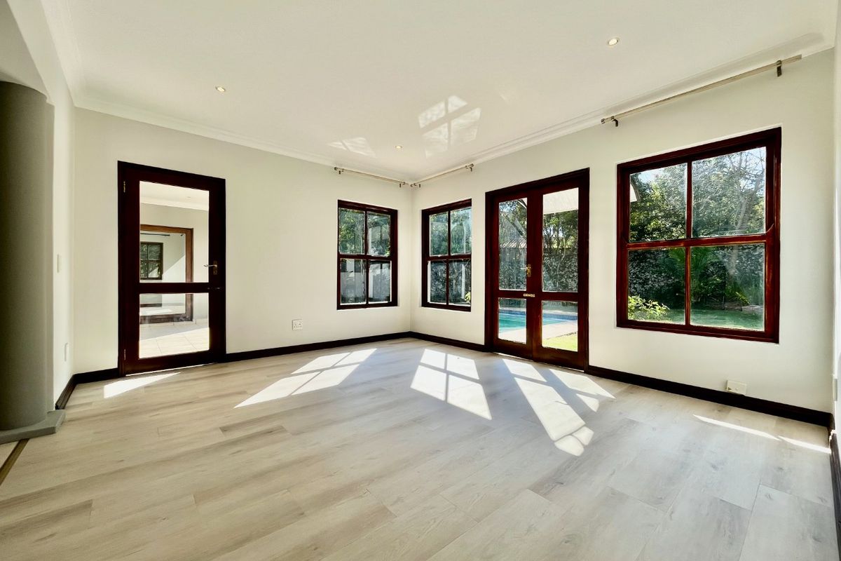 A bright, empty living room featuring light laminate flooring, timber-framed windows, and glass doors providing views of the garden and pool area.