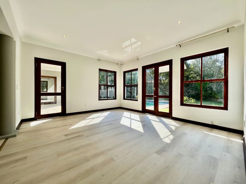 A bright, empty living room featuring light laminate flooring, timber-framed windows, and glass doors providing views of the garden and pool area.