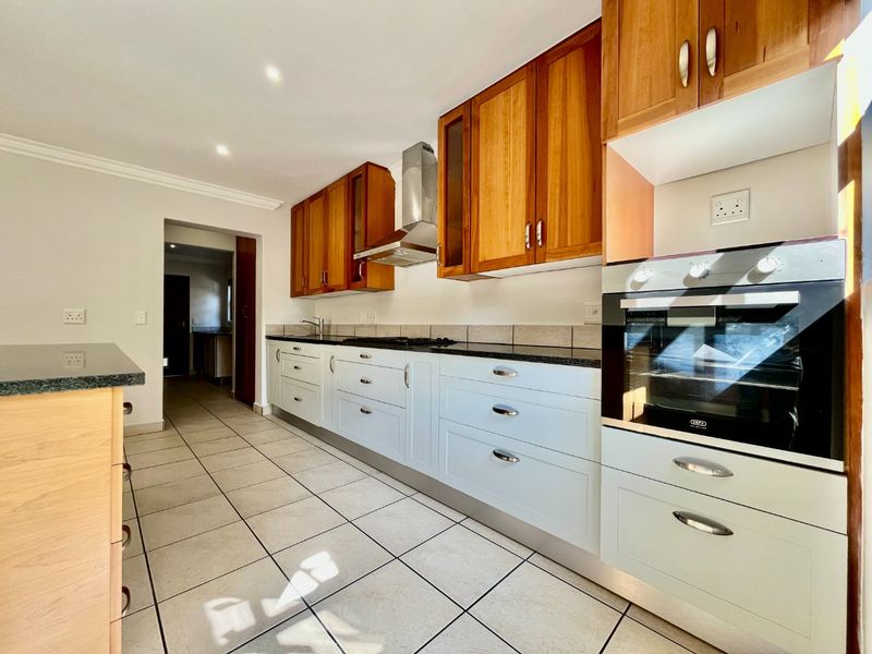 A clean, functional kitchen featuring wooden upper cabinets, light-colored lower cabinetry, granite countertops, built-in oven, and tiled flooring with a view into the scullery.