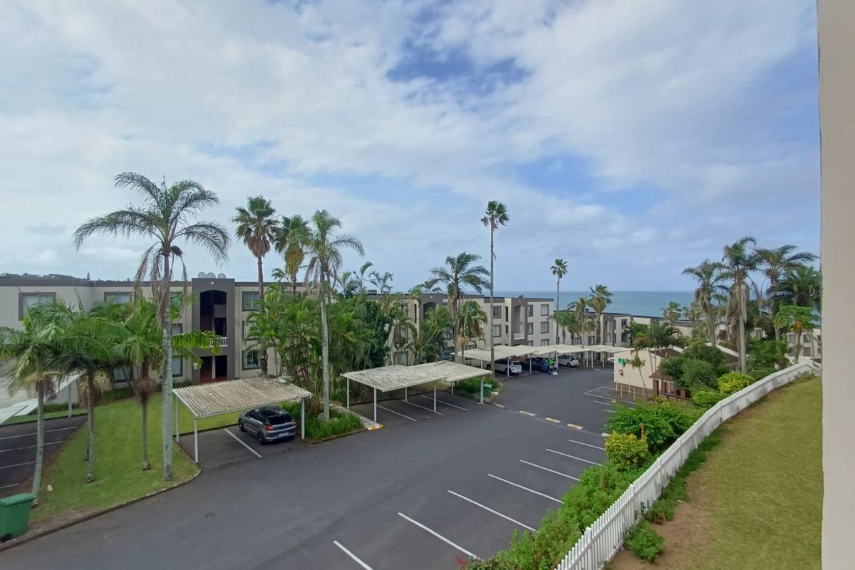 An elevated view of the apartment complex grounds, featuring paved parking bays with carports, landscaped gardens, and distant ocean views.