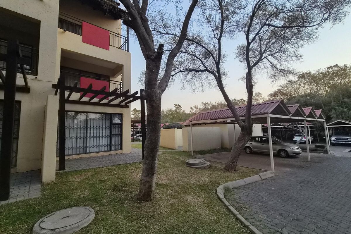 Exterior view of the apartment building featuring a ground floor patio with pergola, upper balconies, and adjacent covered carport parking area.