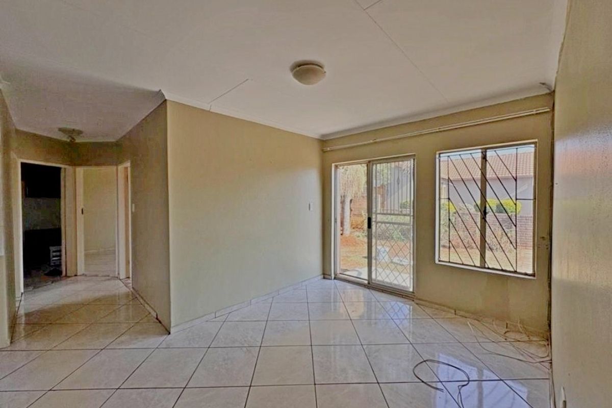 An empty living room featuring tiled flooring, a sliding glass door with security bars, and access to an adjacent hallway.