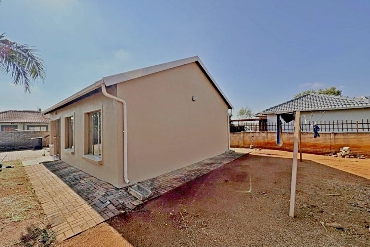 Rear view of the property featuring a paved walkway, exterior wall with guttering, and a spacious yard area enclosed by a brick boundary wall.