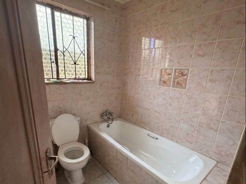 A dated bathroom featuring a standard bathtub, toilet, and full-height wall tiling with a window protected by security bars.
