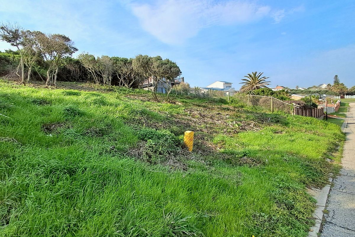 A vacant residential plot featuring a sloped grassy landscape with established trees and boundary fencing, situated along a paved street.