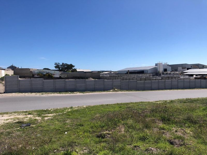 View of an industrial property featuring a precast concrete boundary wall, warehouse structures, and a silo, accessible via an adjacent asphalt road.