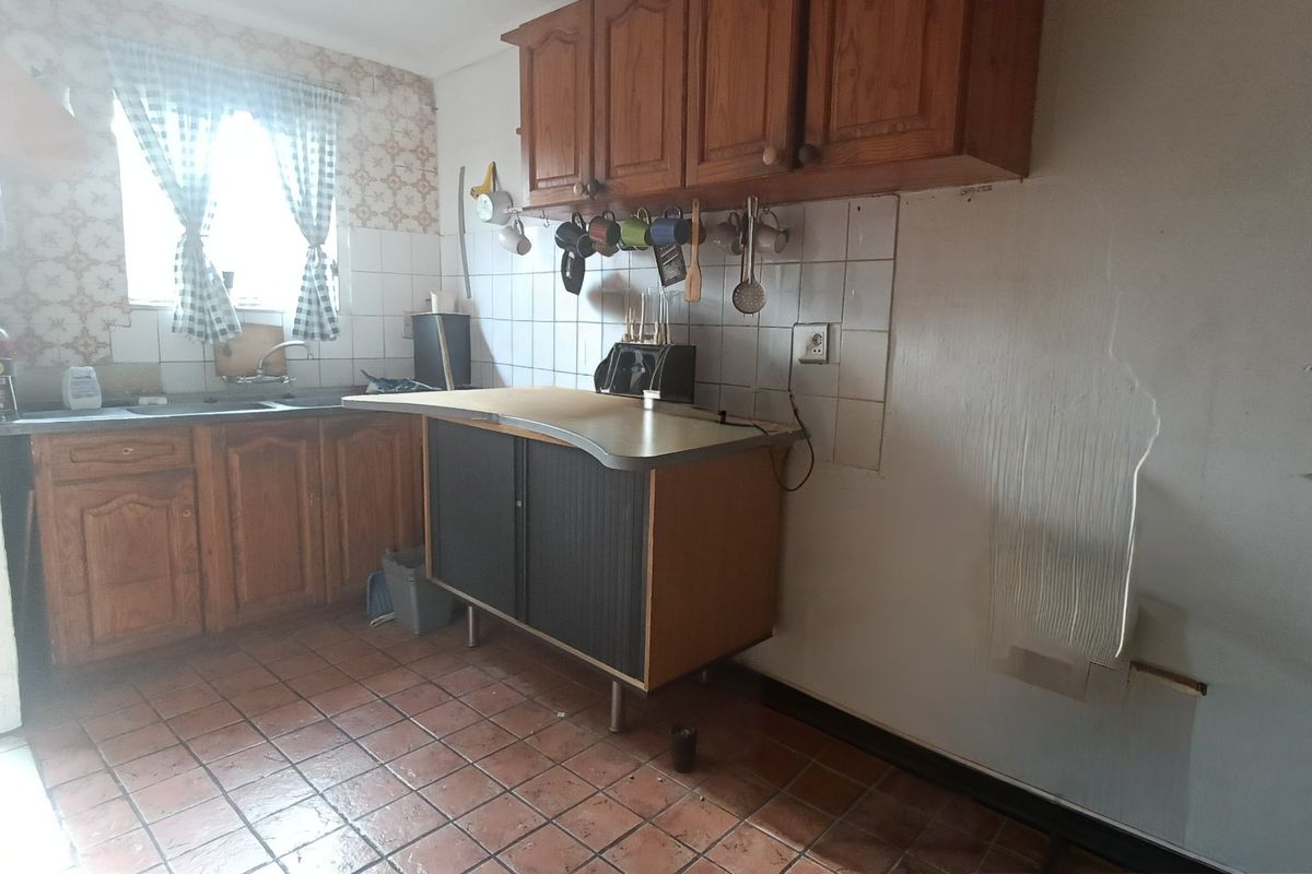 A dated kitchen featuring wooden cabinetry, tiled backsplash, and tiled flooring, currently showing signs of wear and wall damage.