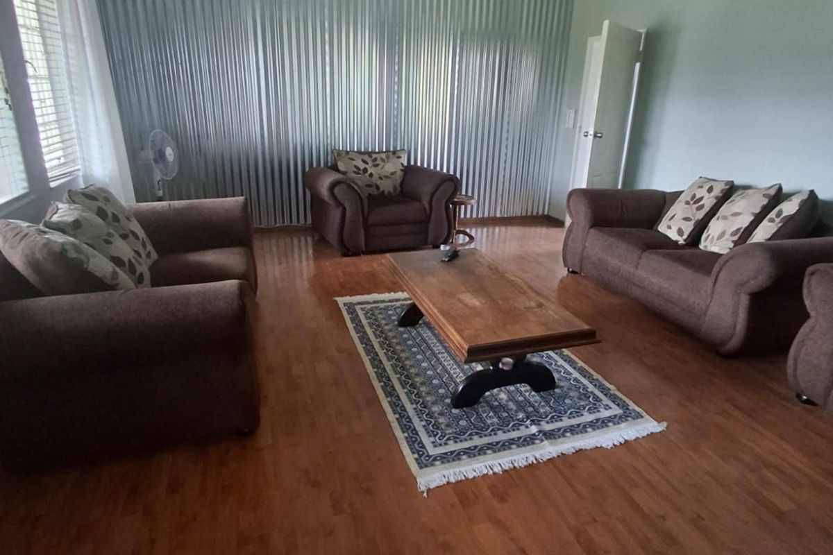 A spacious living room featuring laminate flooring, a decorative ceiling rose with a chandelier, and full-length vertical blinds.