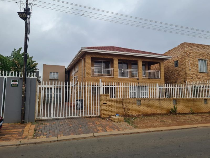 Front view of a two-story face brick house featuring a balcony, secure metal fencing, and a paved driveway.