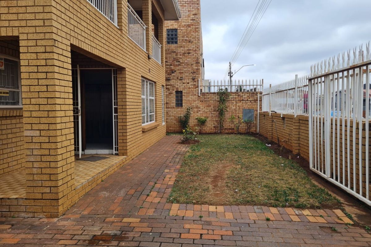 A rear exterior view featuring a face brick house, a paved patio area, a small lawn, and secure perimeter fencing.