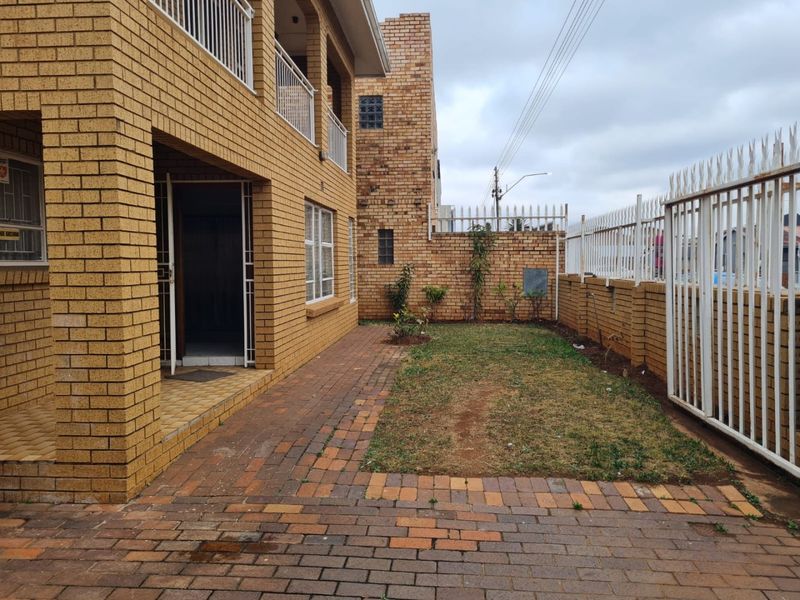 A rear exterior view featuring a face brick house, a paved patio area, a small lawn, and secure perimeter fencing.