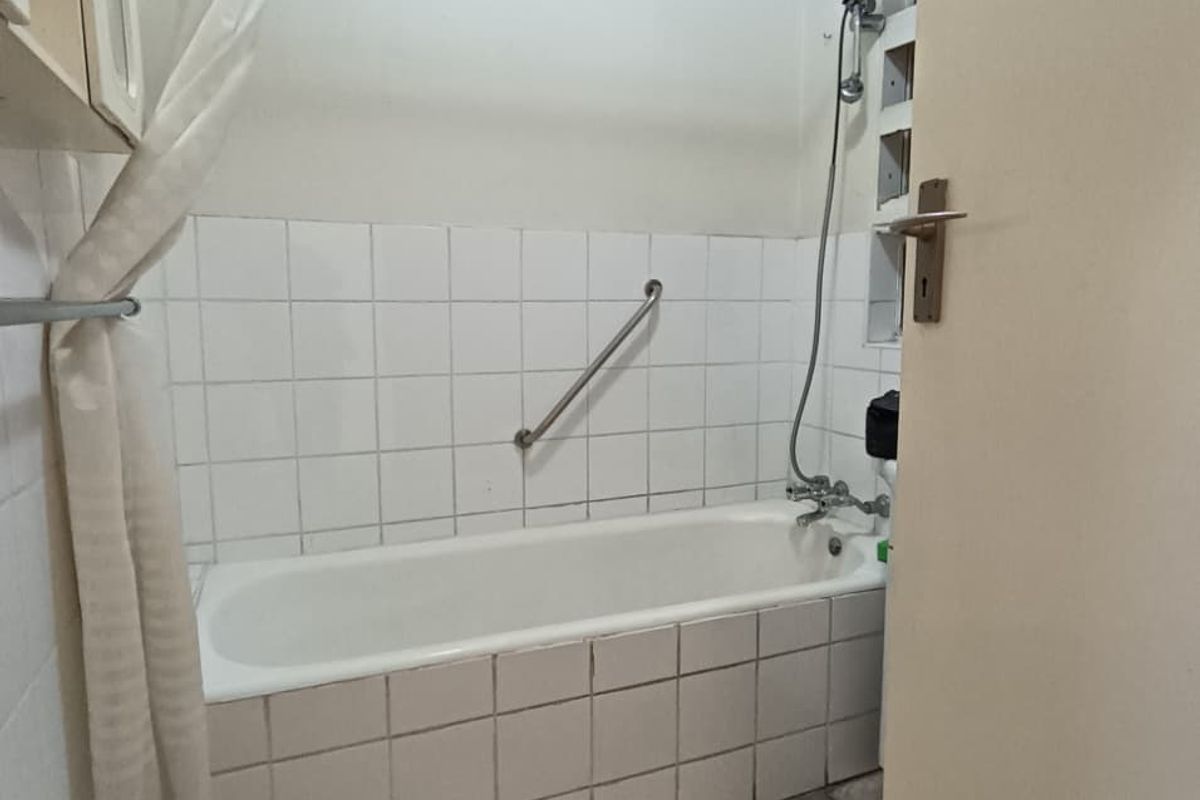 A functional bathroom featuring a bathtub with a shower mixer, grab rail, and tiled finishes, currently requiring cosmetic updates and maintenance.