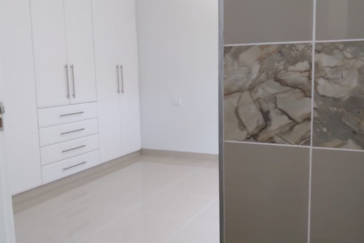 A bright, empty bedroom featuring modern white built-in cupboards, neutral tiled flooring, and a decorative tiled accent wall.