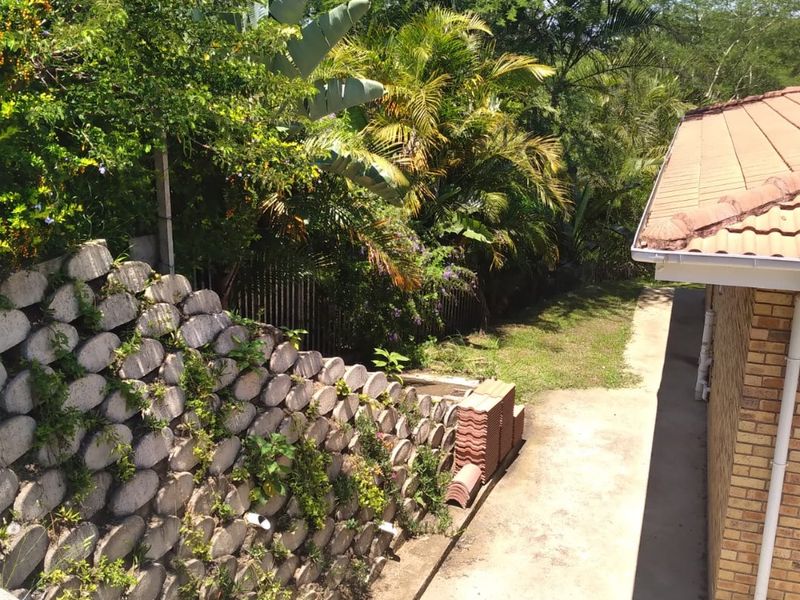 Rear exterior view of the townhouse featuring a brick facade, tiled roof, concrete pathway, and a tiered retaining wall with established garden greenery.