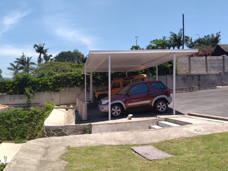 An open-air carport structure providing covered parking, situated on a concrete driveway with adjacent retaining walls and landscaped grounds.