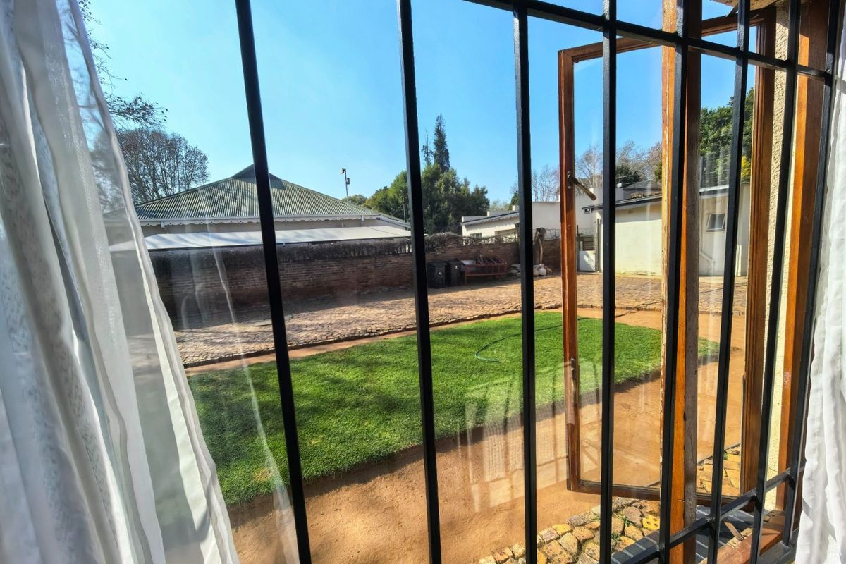 View of the rear garden featuring a lawn area, cobblestone paving, and a brick boundary wall, viewed through a window with security bars.