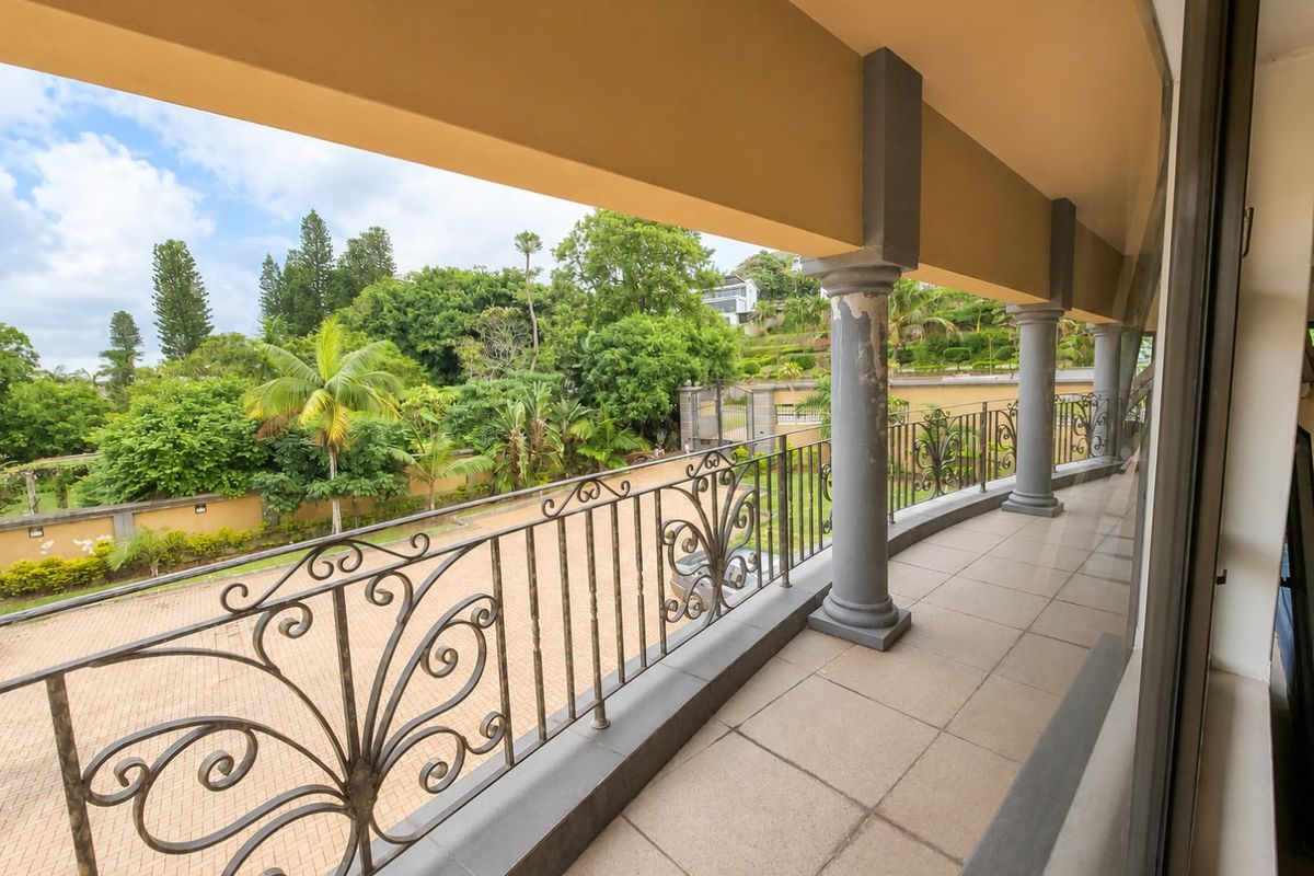 A spacious covered balcony featuring tiled flooring, decorative wrought iron railings, and structural columns overlooking a landscaped garden area.