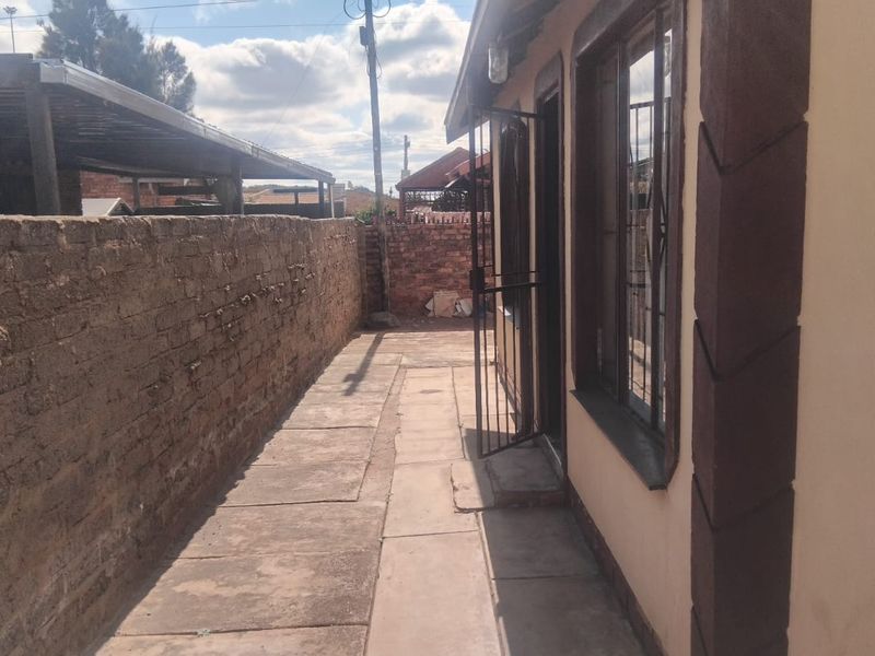 A side exterior view of the property featuring a paved walkway, brick boundary wall, and security gate access.