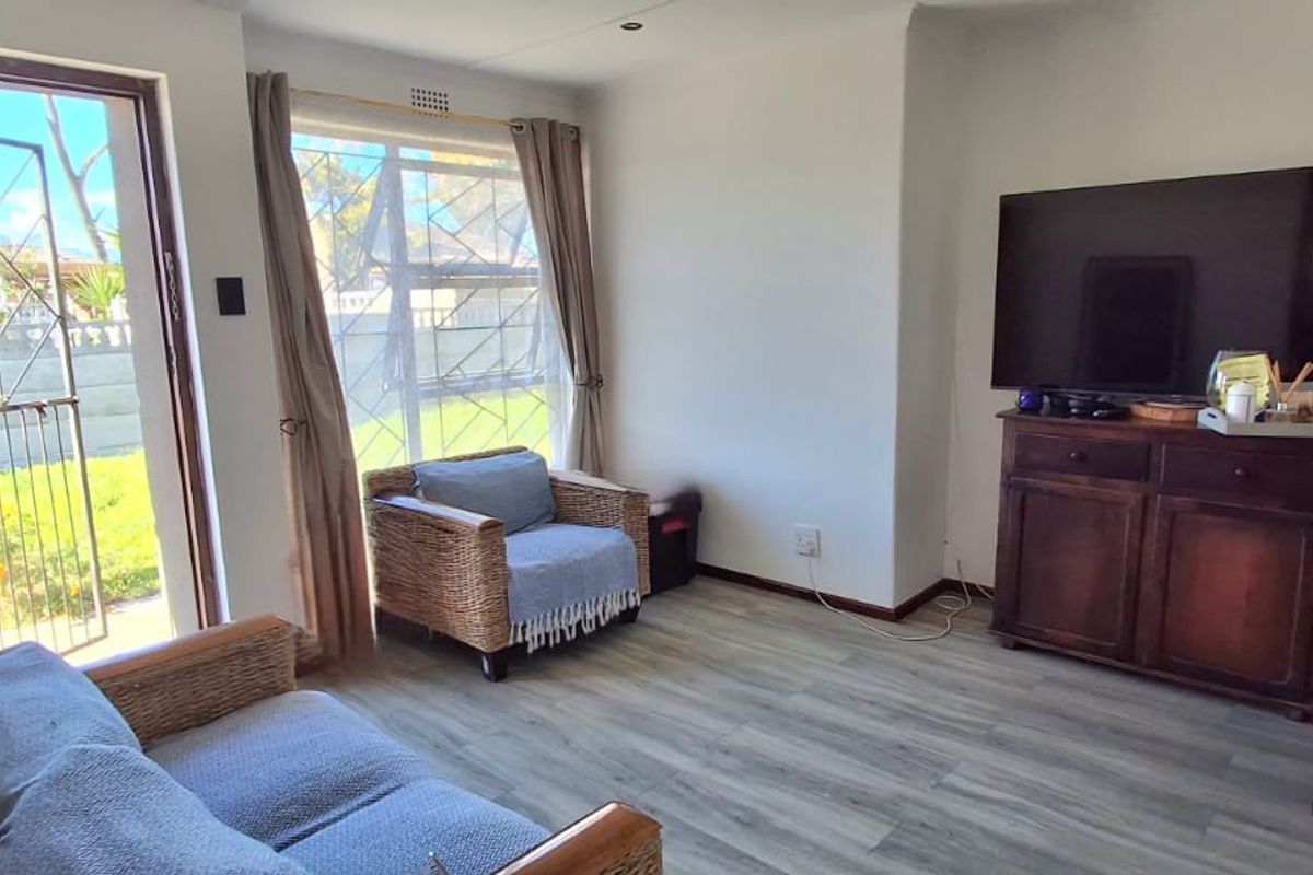 A bright living room featuring modern laminate flooring, large windows with natural light, and direct access to the exterior via a security gate.