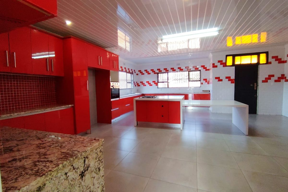 Spacious kitchen featuring vibrant red cabinetry, granite countertops, a central island with a built-in hob, and tiled flooring throughout.