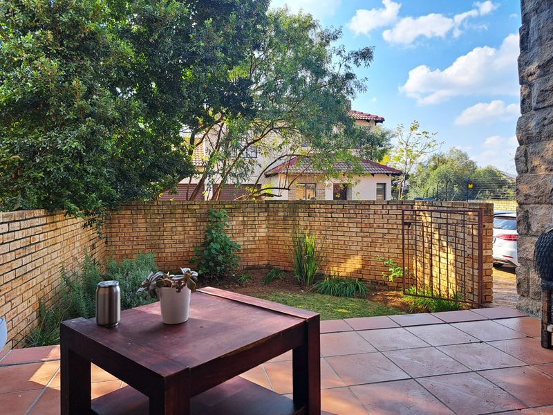 A private outdoor patio area featuring tiled flooring, enclosed by a brick boundary wall with a metal gate, overlooking a small garden space.