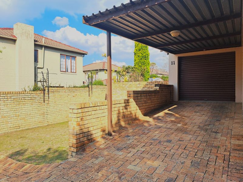 Front exterior view of a townhouse featuring a paved driveway, covered carport, and a secure roll-up garage door with brick boundary walling.