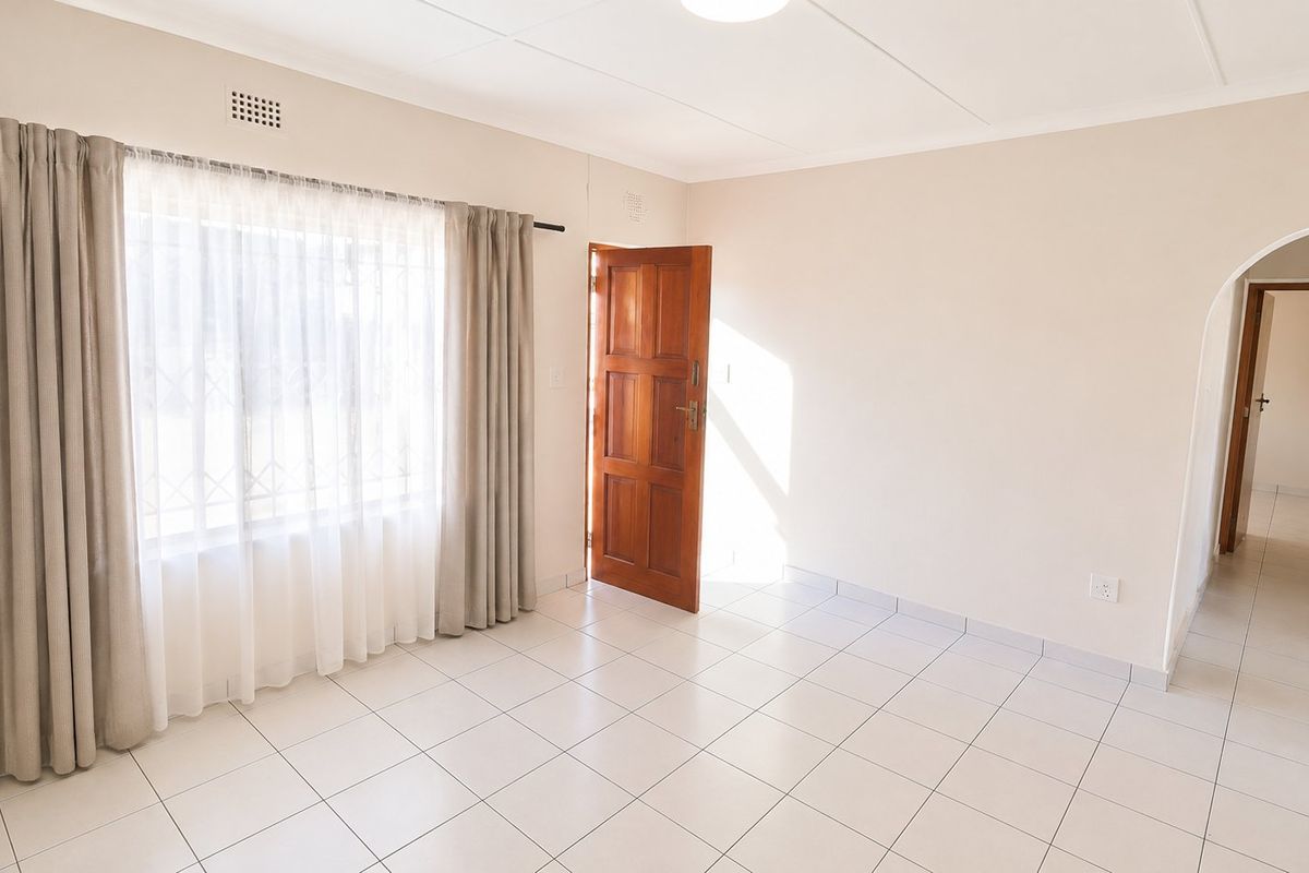 A bright, empty living room featuring neutral tiled flooring, a timber entrance door, and an arched doorway leading to a hallway.