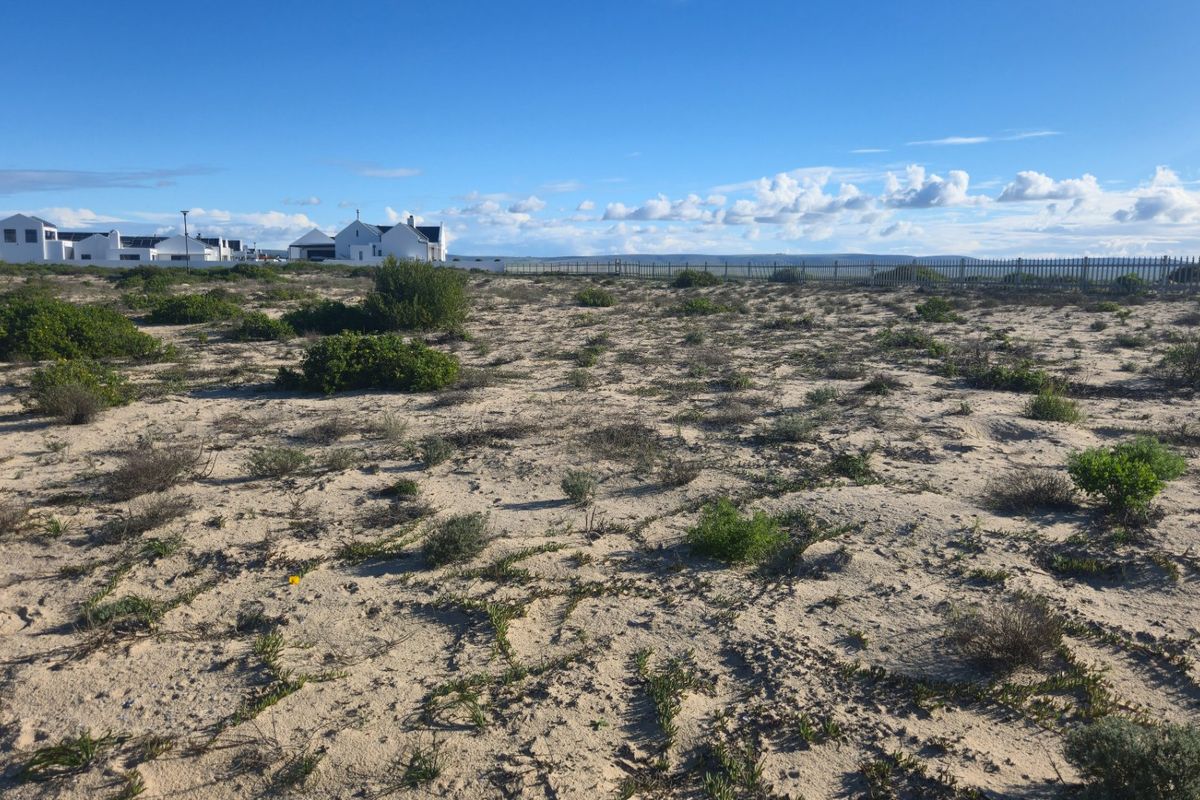 A vacant residential plot featuring sandy terrain, native coastal vegetation, and perimeter fencing, situated in a developed residential estate.