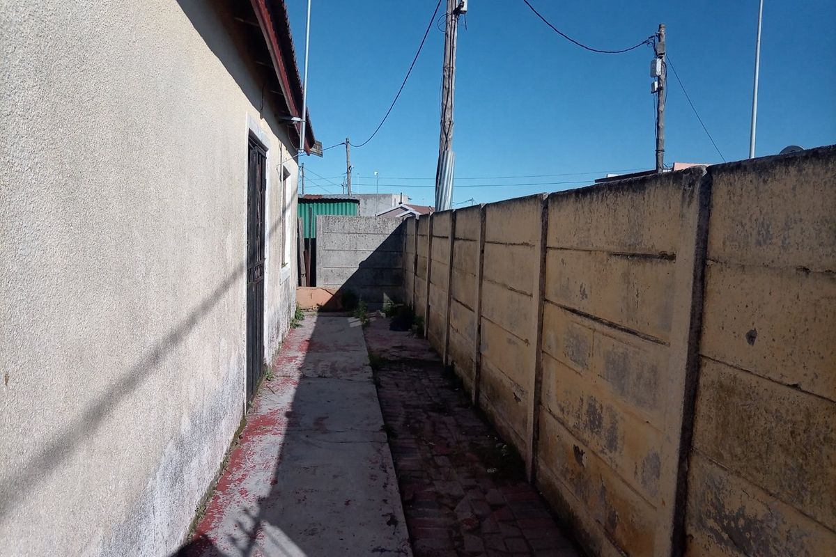 A narrow side passage featuring a concrete pathway, security gate, and a precast concrete boundary wall showing signs of weathering.