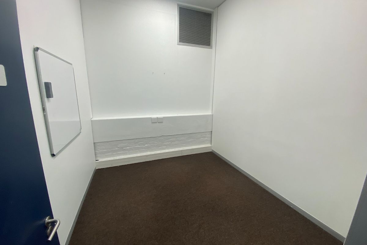 A small, empty office space featuring neutral white walls, dark carpet flooring, and integrated air conditioning ventilation.